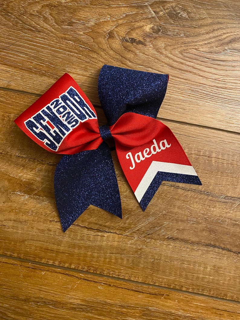 May include: A cheer bow featuring a combination of red fabric and dark blue glitter fabric. The left side displays the word SENIOR in dark blue glitter lettering, while the right side features the name Jaeda in white script with a white chevron accent.