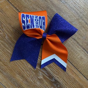 May include: A cheer bow featuring orange and purple glitter fabric. The orange section displays the word SENIOR in blue block letters with a white outline. The bow includes long tails with a white chevron stripe detail at the bottom edge.