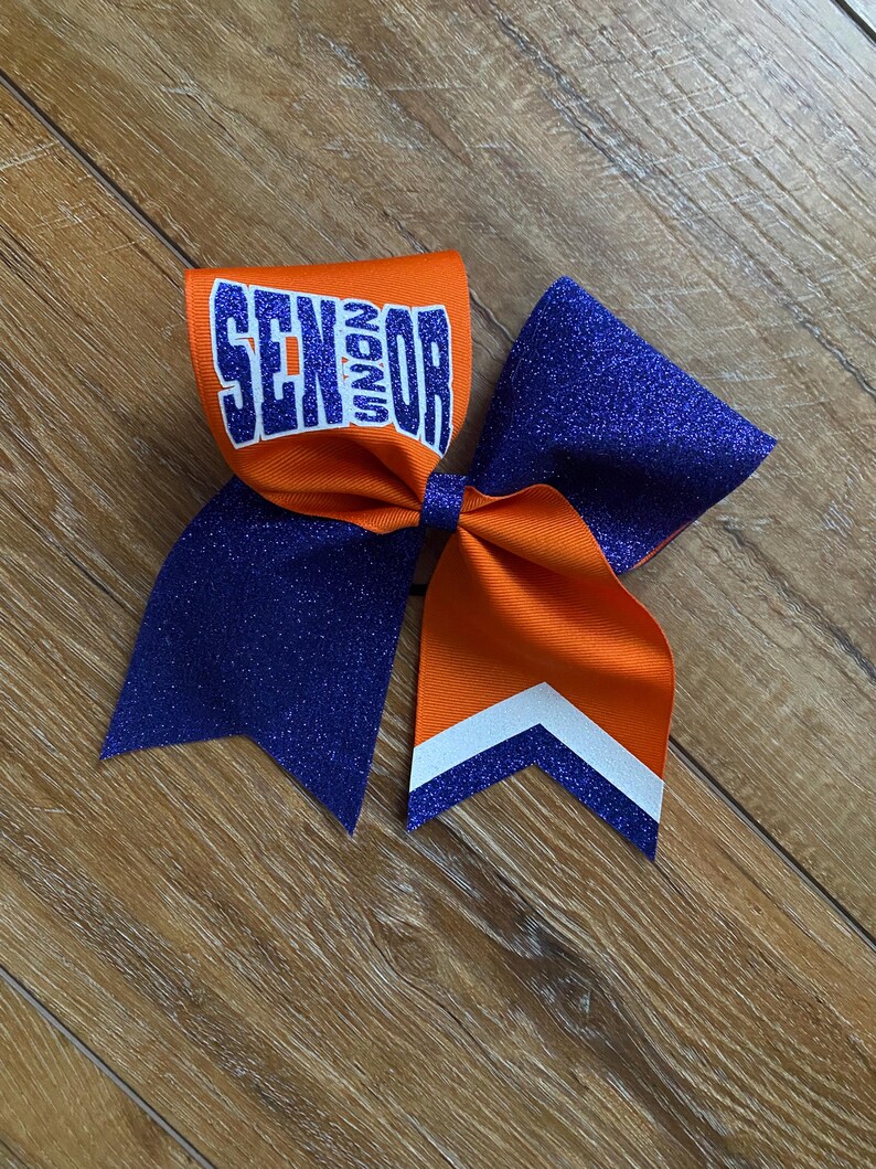 May include: A cheer bow featuring orange and purple glitter fabric. The orange section displays the word SENIOR in blue block letters with a white outline. The bow includes long tails with a white chevron stripe detail at the bottom edge.