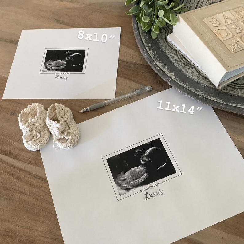 May include: Two white framed sonogram prints, one 8x10 inches and the other 11x14 inches, with the text "Wishes for Lucas". A pair of cream-colored crocheted baby booties sits next to the prints. A pen is also visible.