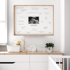 May include: A framed baby shower guest book with a sonogram image and the text "Wishes for Baby Smith." The frame is light brown, and the background is white with handwritten messages. The guest book is displayed on a white dresser.