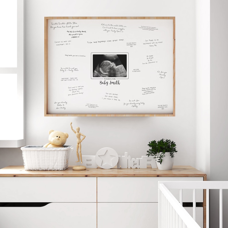 May include: A framed baby shower guest book with a sonogram image and the text "Wishes for Baby Smith." The frame is light brown, and the background is white with handwritten messages. The guest book is displayed on a white dresser.