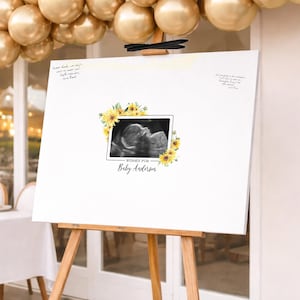 May include: A white rectangular sign on a wooden easel featuring a central ultrasound image framed by yellow sunflowers. The text reads "WISHES FOR Baby Anderson" with handwritten notes in the top corners. The sign is displayed beneath gold balloons.