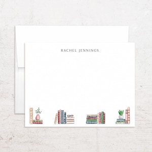 May include: A white stationery card with the name RACHEL JENNINGS printed in black at the top center. The bottom border features a colorful watercolor illustration of various stacks of books, small potted plants, and a pair of eyeglasses.