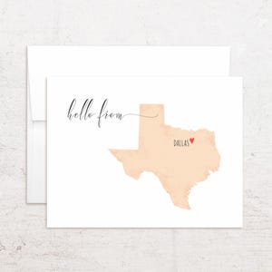 May include: A white card with a peach-colored Texas map and the word "hello from" in elegant script. The city of Dallas is marked with a red heart. The card is inside a white envelope.
