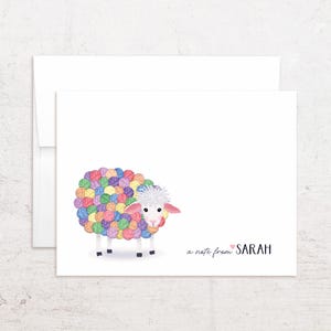 May include: A white stationery card and envelope featuring a cartoon sheep with a body made of colorful yarn balls. The sheep has a white face, pink ears, and a tuft of white hair. The text reads a note from SARAH with a small pink heart.