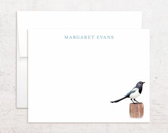 May include: A white stationery card and matching envelope featuring a black and white magpie bird perched on a brown wooden post in the lower right corner. The name MARGARET EVANS is printed in light blue capital letters at the top center.