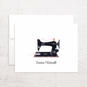 May include: A white folded greeting card featuring a black vintage sewing machine illustration with a pink thread spool and a thin pink thread line. The name Diane Mitchell is printed in black text below the sewing machine graphic.