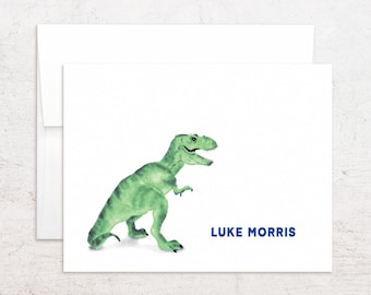 May include: A white folded greeting card featuring a watercolor illustration of a green Tyrannosaurus Rex dinosaur on the left side and the name LUKE MORRIS printed in bold blue capital letters on the bottom right side, paired with a white envelope.