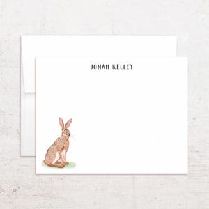 May include: A white stationery card and matching envelope featuring a watercolor illustration of a brown hare sitting on a small patch of green grass in the lower left corner. The name JONAH KELLEY is printed in black sans-serif text at the top center.