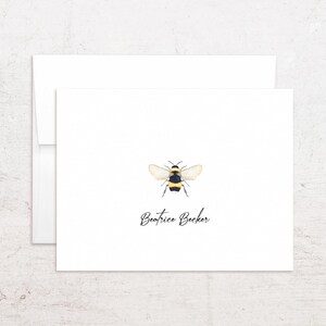 May include: White stationery set featuring a watercolor illustration of a bee. The bee is black and yellow with translucent wings. The name "Beatrice Becker" is written in cursive below the bee. Includes a matching envelope.