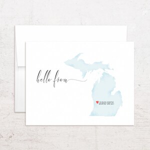 May include: A white card with a light blue watercolor map of Michigan, with a red heart marking Grand Rapids. The words "hello from" are written in elegant script. An envelope is behind the card.