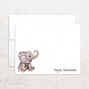 May include: White stationery set featuring a watercolor illustration of a gray baby elephant. The elephant has a raised trunk and is sitting down. The name "Hazel Stevenson" is written in black script on the bottom right corner. Includes a white envelope.