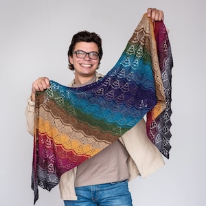 Puede incluir: Una persona sosteniendo una chal de ganchillo de colores arcoíris con un patrón de encaje. El chal está hecho de hilo en tonos de gris, morado, azul, verde, amarillo, naranja y rojo.