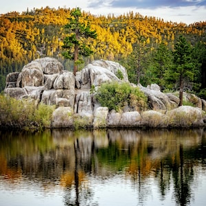 May include: A scenic landscape featuring a calm lake reflecting the surrounding trees and rocks. The foreground shows the dark water, with a rocky island in the middle. The background features a forest with golden and green trees under a cloudy sky.