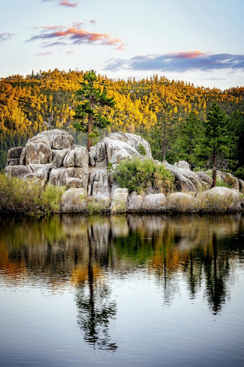 May include: A scenic landscape featuring a calm lake reflecting the surrounding trees and rocks. The foreground shows the dark water, with a rocky island in the middle. The background features a forest with golden and green trees under a cloudy sky.
