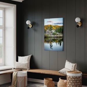 May include: A vertical canvas print of a serene lake scene with a reflection of trees and rocks. The artwork is displayed on a dark gray wall, complemented by a wooden bench, decorative baskets, and pillows, creating a cozy interior design.