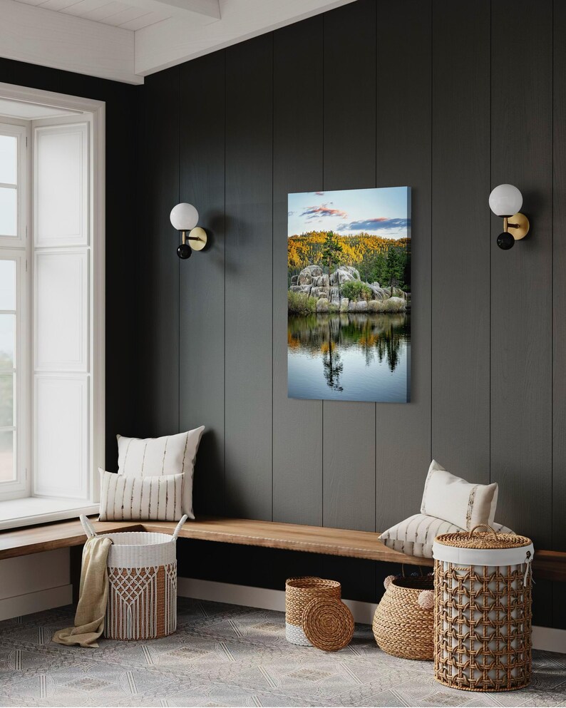 May include: A vertical canvas print of a serene lake scene with a reflection of trees and rocks. The artwork is displayed on a dark gray wall, complemented by a wooden bench, decorative baskets, and pillows, creating a cozy interior design.