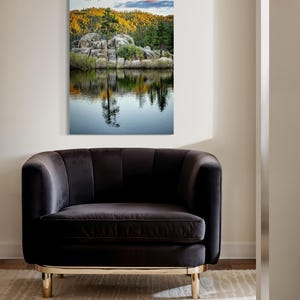 May include: A framed landscape print depicts a serene lake scene with reflections of trees and rocks. The artwork hangs above a dark gray velvet armchair with gold-colored legs, set on a patterned rug. The scene captures a tranquil outdoor setting.
