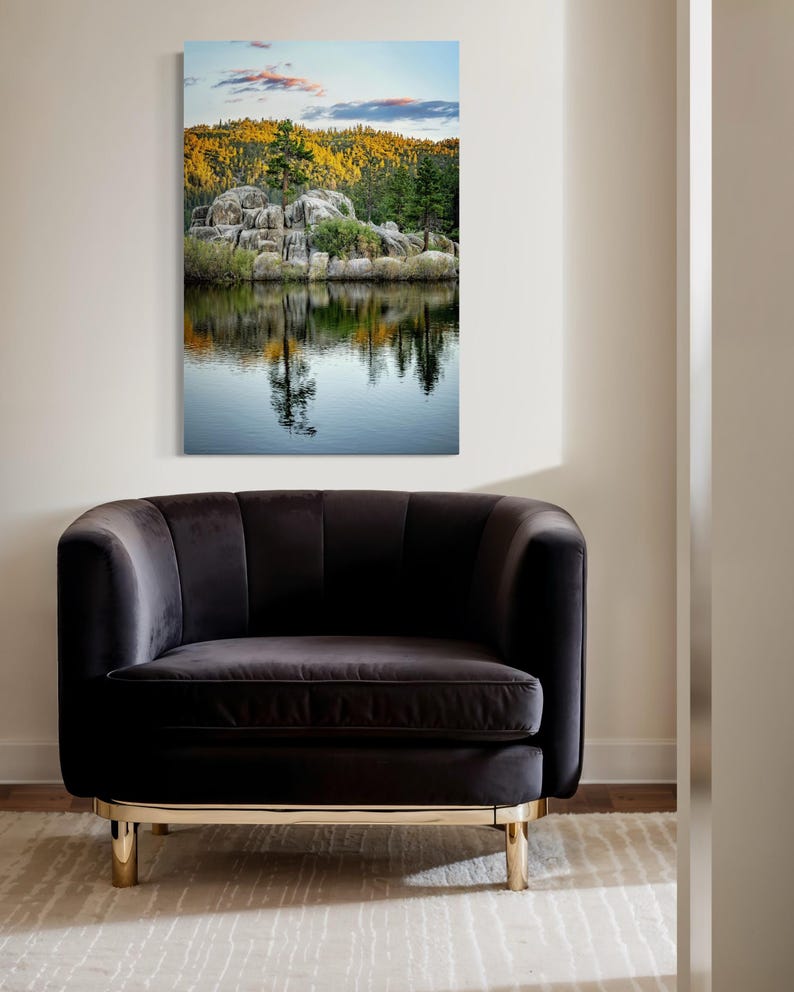May include: A framed landscape print depicts a serene lake scene with reflections of trees and rocks. The artwork hangs above a dark gray velvet armchair with gold-colored legs, set on a patterned rug. The scene captures a tranquil outdoor setting.