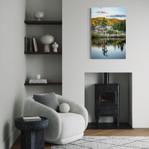 May include: A modern living room with a white armchair, a dark blue side table, and a gray rug. A landscape painting of a lake and trees hangs on the wall. A black fireplace and shelves with decor complete the scene.