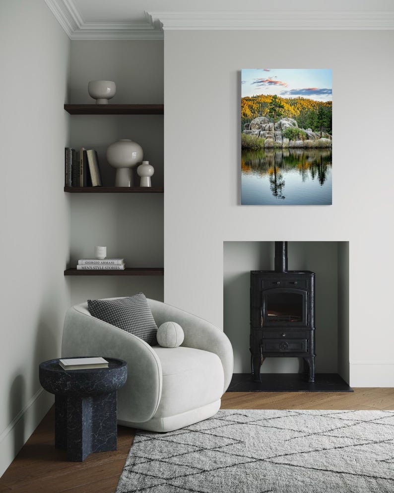 May include: A modern living room with a white armchair, a dark blue side table, and a gray rug. A landscape painting of a lake and trees hangs on the wall. A black fireplace and shelves with decor complete the scene.
