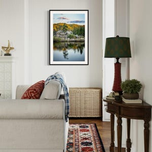 May include: A framed landscape photograph featuring a serene lake reflecting trees and a rocky shoreline. The image is displayed in a living room setting, with a sofa, rug, and side table with a lamp.