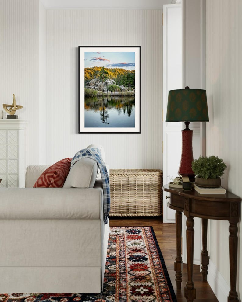 May include: A framed landscape photograph featuring a serene lake reflecting trees and a rocky shoreline. The image is displayed in a living room setting, with a sofa, rug, and side table with a lamp.