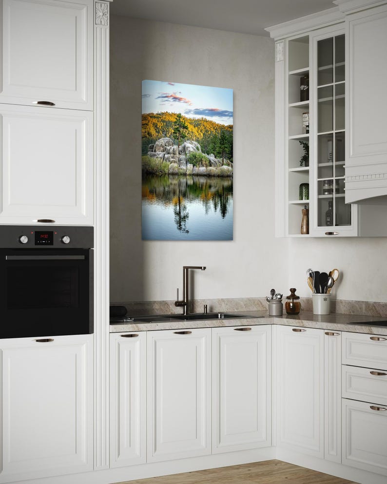 May include: A rectangular landscape canvas print depicting a serene lake scene with a rocky shoreline, lush green trees, and a colorful sunset. The artwork is displayed in a modern white kitchen setting.