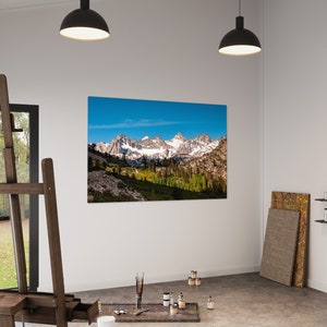 Puede incluir: Una impresi&oacute;n en lienzo de una cordillera con picos cubiertos de nieve y un cielo azul. La impresi&oacute;n est&aacute; colgada en una pared blanca en un estudio de arte. Hay un caballete, pinceles y botes de pintura en el suelo frente a la impresi&oacute;n.