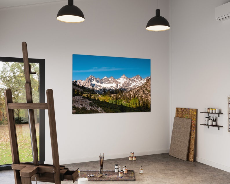 Puede incluir: Una impresi&oacute;n en lienzo de una cordillera con picos cubiertos de nieve y un cielo azul. La impresi&oacute;n est&aacute; colgada en una pared blanca en un estudio de arte. Hay un caballete, pinceles y botes de pintura en el suelo frente a la impresi&oacute;n.