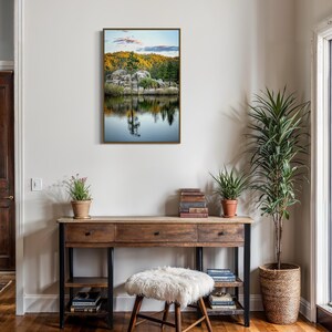 May include: A framed landscape art print featuring a serene lake scene with reflections of trees and rocks. The artwork hangs above a wooden desk with drawers, a fluffy stool, and a patterned rug. A tall potted plant and other decor complete the interior.