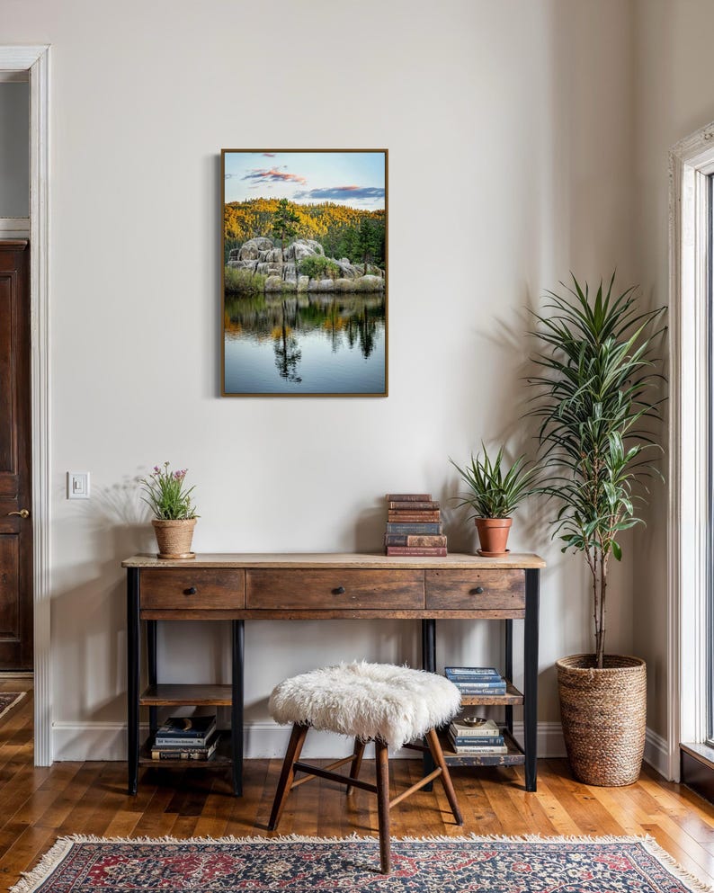 May include: A framed landscape art print featuring a serene lake scene with reflections of trees and rocks. The artwork hangs above a wooden desk with drawers, a fluffy stool, and a patterned rug. A tall potted plant and other decor complete the interior.