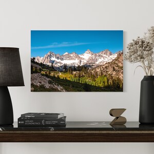 Puede incluir: Una fotograf&iacute;a panor&aacute;mica de una cordillera con picos cubiertos de nieve y un valle con &aacute;rboles verdes en primer plano. El cielo es azul y despejado.