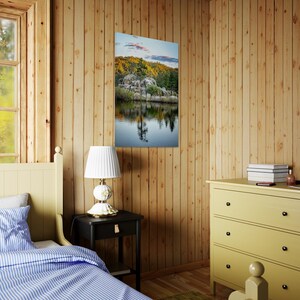 May include: A framed landscape print depicting a serene lake scene with rocky formations, trees, and a reflection in the water. The print hangs on a wooden wall in a bedroom setting, alongside a nightstand, lamp, and dresser.
