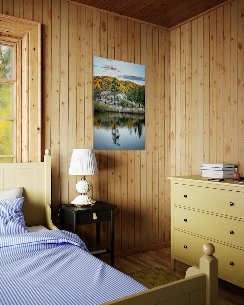 May include: A framed landscape print depicting a serene lake scene with rocky formations, trees, and a reflection in the water. The print hangs on a wooden wall in a bedroom setting, alongside a nightstand, lamp, and dresser.