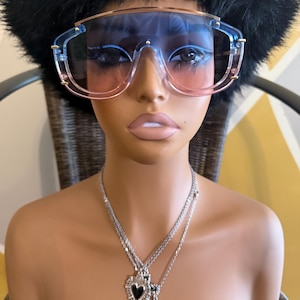 May include: Oversized shield sunglasses with a gradient blue-to-pink lens and a gold-tone top bar. The eyewear is paired with a layered silver-tone chain necklace featuring two heart-shaped pendants, one with a black inlay and one with a cross.