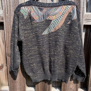 May include: A black long-sleeve sweater with gold metallic flecks throughout. The upper back features an intricate appliqué design with grey, brown, and multi-colored paisley fabric panels, decorative cutouts, and small metallic studs.