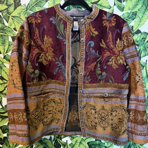 May include: A long-sleeve jacket featuring a tapestry-style floral and scroll pattern in shades of burgundy, gold, and lavender. The jacket has a round neckline, a front button closure with silver-toned metal buttons, and two front pockets.