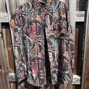 May include: A long-sleeve button-down shirt featuring a dark base color with an intricate pattern of interlocking geometric shapes and abstract motifs in shades of pink, teal, and tan. The shirt includes a standard collar and a single chest pocket.