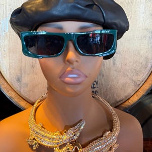 May include: A mannequin head wearing a black leather beret and dark green sunglasses. A gold-colored necklace with two bejeweled alligator pendants is around the neck. The background is a wooden barrel.
