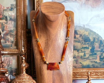 Op de afbeelding: Een ketting met een grote, oranje carneol hanger en bruine en groene kralen. De ketting wordt getoond op een houten mannequin tegen een achtergrond van ingelijste kunstwerken.