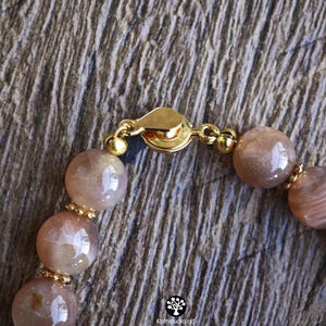 May include: A beaded bracelet featuring smooth, round, light brown sunstone beads with a subtle shimmer. The beads are separated by small, gold-toned metal spacers and secured with a gold-toned circular clasp. The text Kathstudio.art is visible.