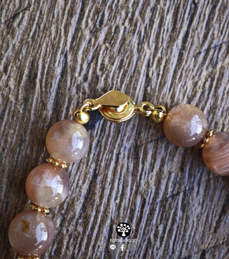 May include: A beaded bracelet featuring smooth, round, light brown sunstone beads with a subtle shimmer. The beads are separated by small, gold-toned metal spacers and secured with a gold-toned circular clasp. The text Kathstudio.art is visible.