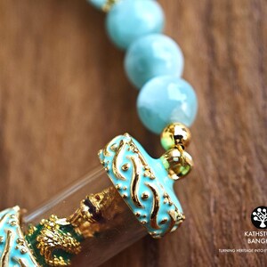 K&ouml;nnte beinhalten: Ein Armband mit hellblauen, kugelf&ouml;rmigen Perlen und einem zentralen, zylindrischen Anh&auml;nger aus klarem Glas. Der Anh&auml;nger ist mit goldfarbenem Metall mit t&uuml;rkisfarbenen Emaille-Verzierungen versehen und enth&auml;lt eine kleine goldfarbene Figur.
