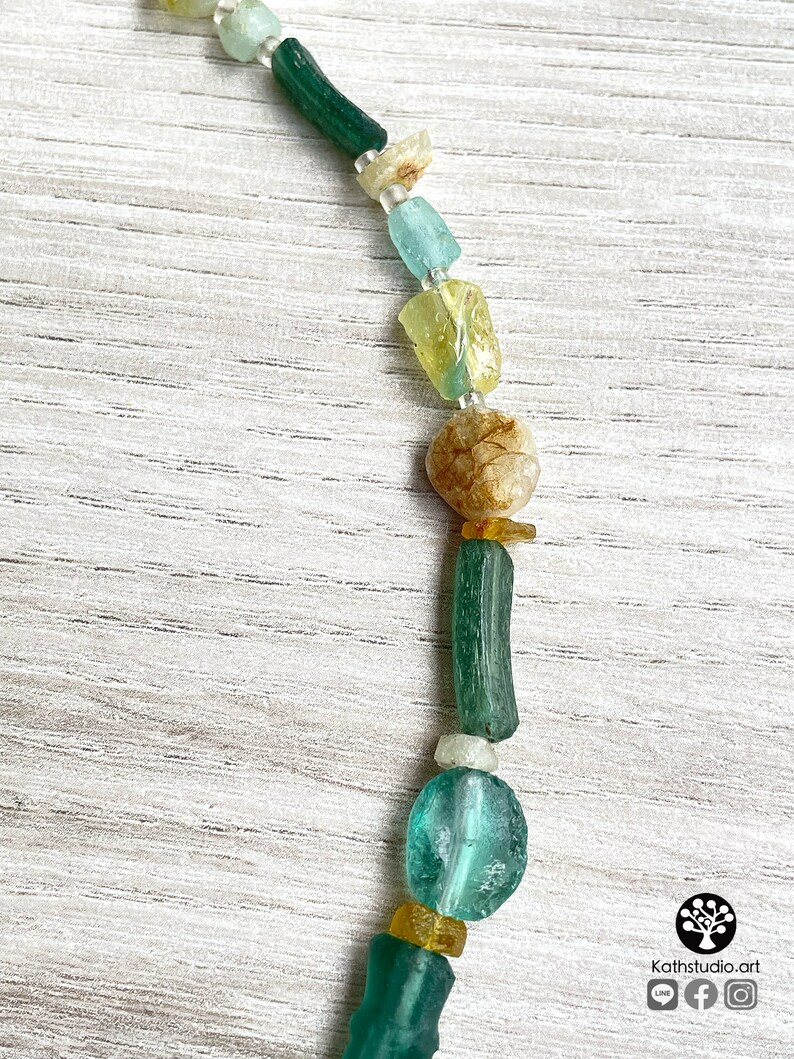 Puede incluir: Primer plano de un collar con una variedad de cuentas coloridas de forma irregular. Las cuentas son en tonos verdes, azules, amarillos y beige. El collar se muestra sobre una superficie de madera de color claro. El logotipo del artista est&aacute; en la esquina inferior derecha.