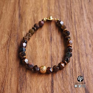 May include: A beaded bracelet featuring faceted brown and dark gray tiger eye stones arranged in a circular pattern. The bracelet includes a single central gold-toned spherical bead with a detailed floral engraving and a gold-toned metal clasp.