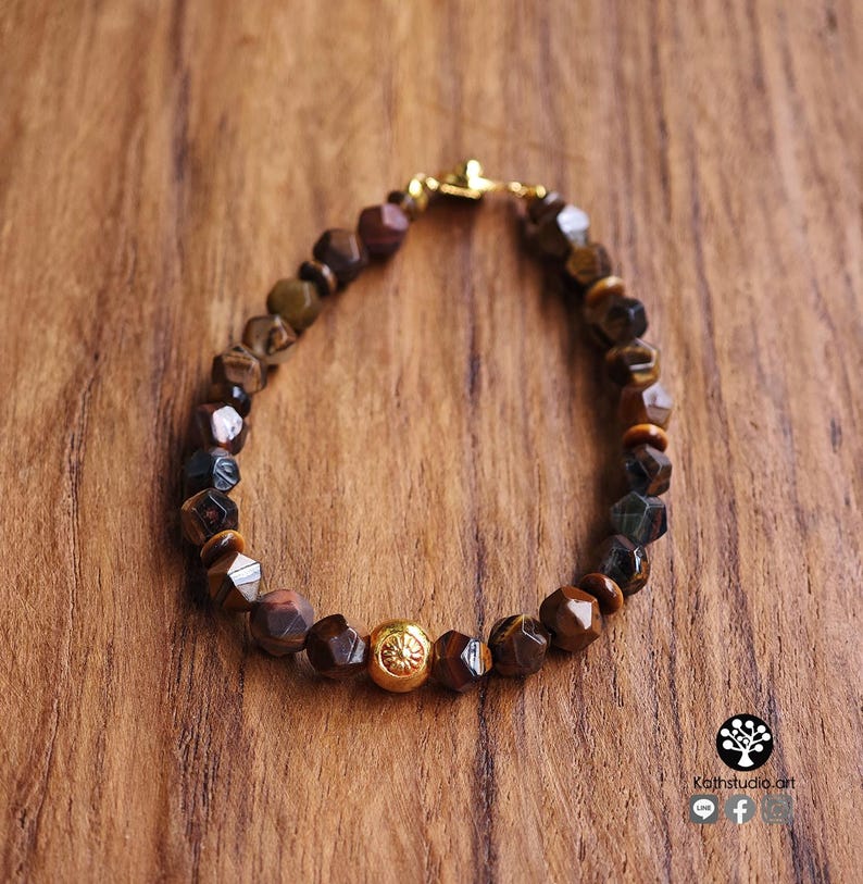 May include: A beaded bracelet featuring faceted brown and dark gray tiger eye stones arranged in a circular pattern. The bracelet includes a single central gold-toned spherical bead with a detailed floral engraving and a gold-toned metal clasp.