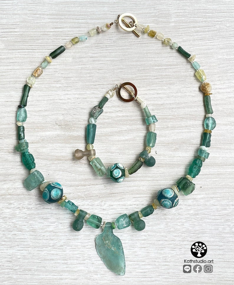 Puede incluir: Un conjunto de collar y pulsera con cuentas de vidrio verdes y azules. El collar tiene un colgante. La pulsera y el collar tienen un cierre de palanca. El conjunto se exhibe sobre una superficie de madera clara.