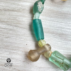 Puede incluir: Primer plano de un collar con cuentas de vidrio marino de varias formas y tama&ntilde;os, en tonos verdes, azules y amarillos. Las cuentas est&aacute;n ensartadas, creando un dise&ntilde;o &uacute;nico y org&aacute;nico. El collar est&aacute; sobre una superficie de madera clara.